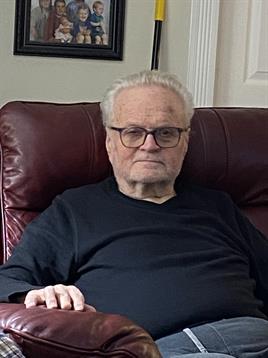 An elderly man with glasses sits in a comfortable leather chair, appearing thoughtful and relaxed.