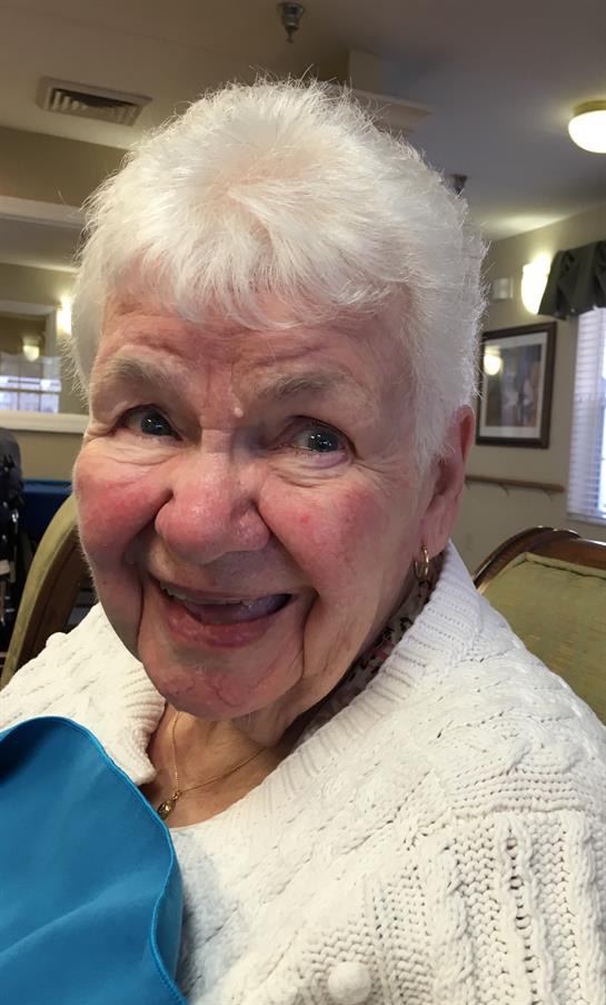 Elderly woman with short white hair smiles happily while seated in a warm indoor setting.
