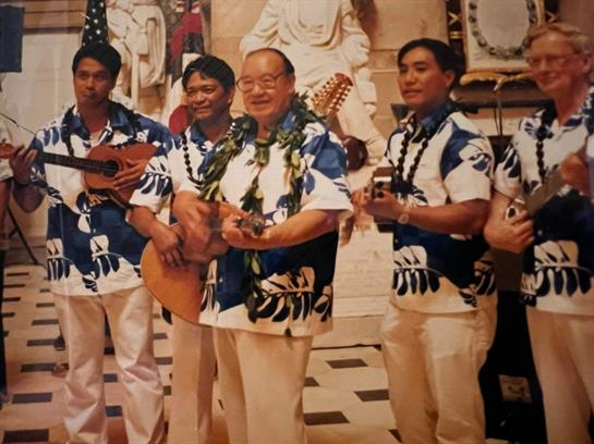 Five musicians in floral shirts play Hawaiian instruments at a cultural celebration.