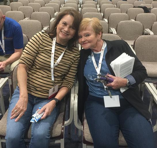 Two women sit close together, smiling and enjoying their time at a conference in an auditorium.