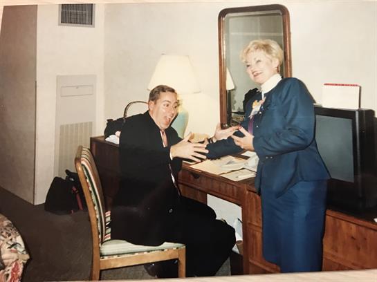 A man and a woman engage in conversation at a desk in a hotel room, both appearing professional.