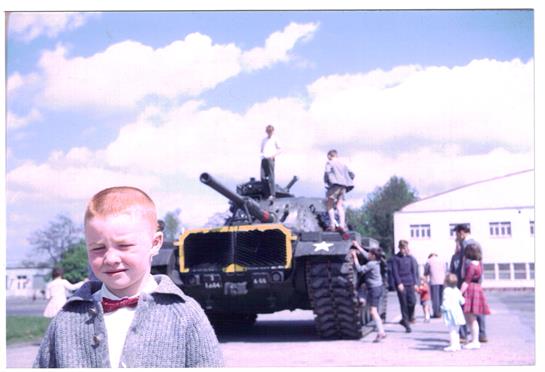 A young boy stands in front of a military tank while various individuals gather around celebrating.