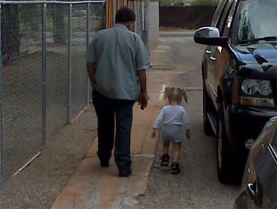 A man walks hand in hand with a small child down a narrow pathway beside parked vehicles.