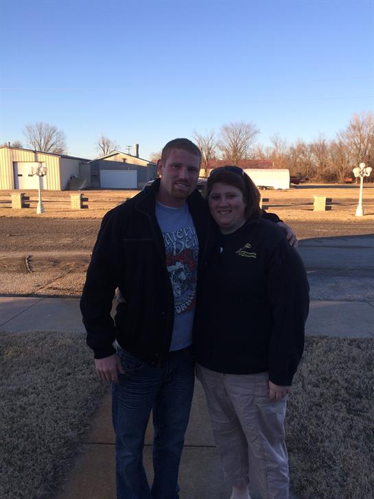 A smiling couple stands on a pathway against a backdrop of buildings and trees in winter.