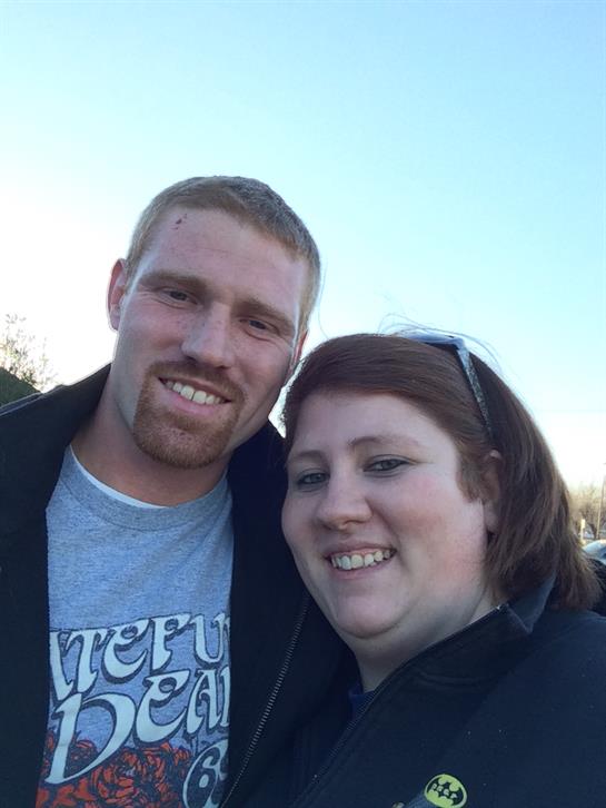 A couple stands closely together, both smiling against a clear blue sky, enjoying their time.