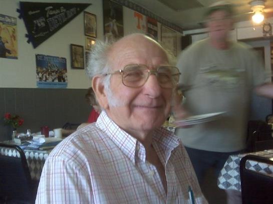 A cheerful elderly man is seated at a table in a warm diner, engaging with people nearby.