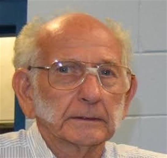 An elderly man with glasses looks thoughtfully in a classroom environment filled with light.