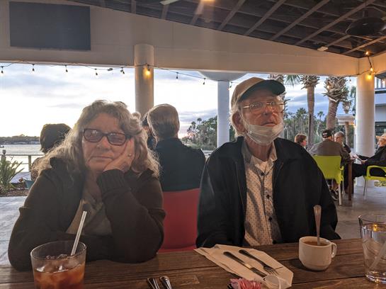 A woman with curly hair and glasses sits thoughtfully beside an elderly man in a cap, both relaxing.