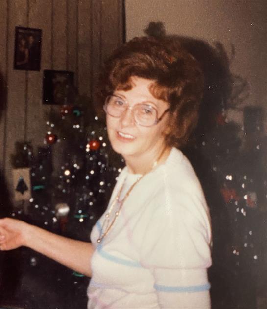 A woman with curly hair and glasses smiles warmly in a festive living room during the holidays.