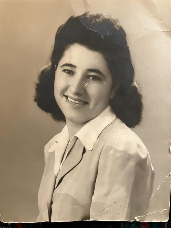 A young woman with dark wavy hair smiles while wearing a formal outfit, exuding a joyful demeanor.