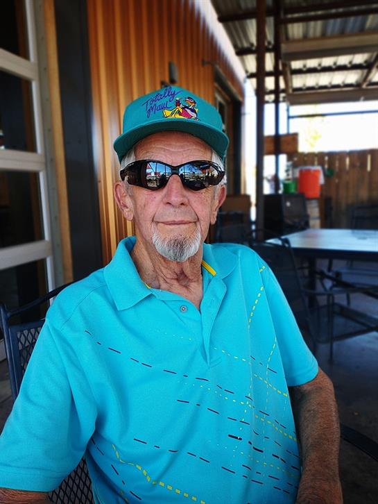 Senior man in a blue shirt and cap relaxes outdoors in a casual dining atmosphere under shade.