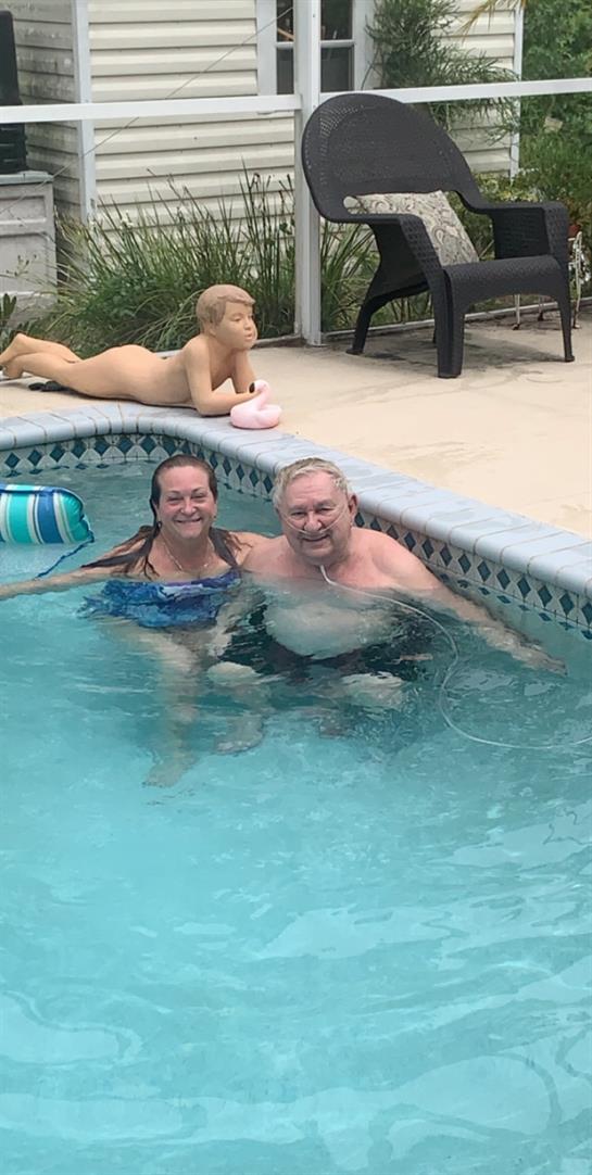 Two people smile as they relax in a pool with playful decor nearby, enjoying a sunny day outdoors.