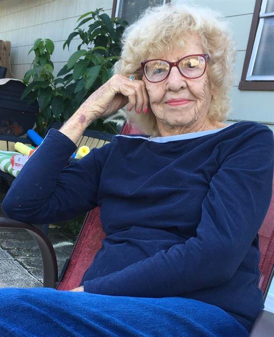An elderly woman relaxes in a chair, enjoying the sunny afternoon and green surroundings.