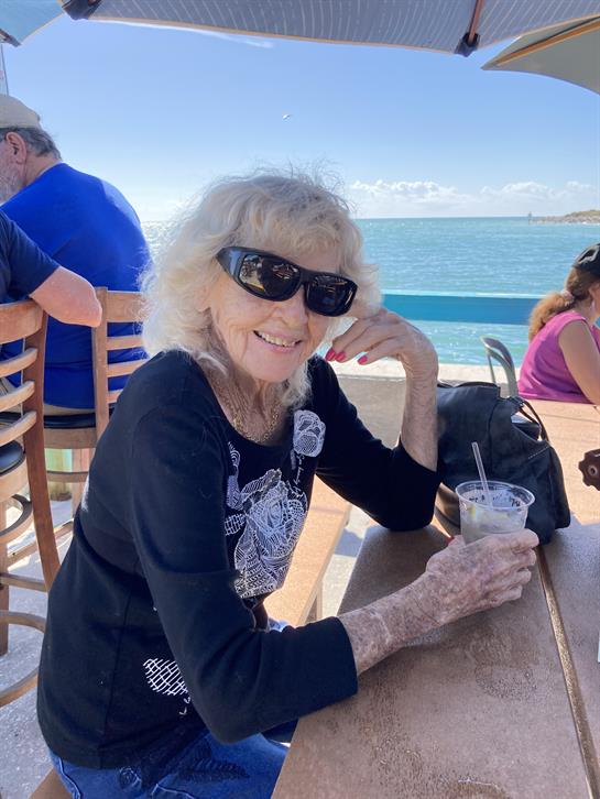A joyful elderly woman sits at a table by the sea, sipping a cold drink and smiling brightly.