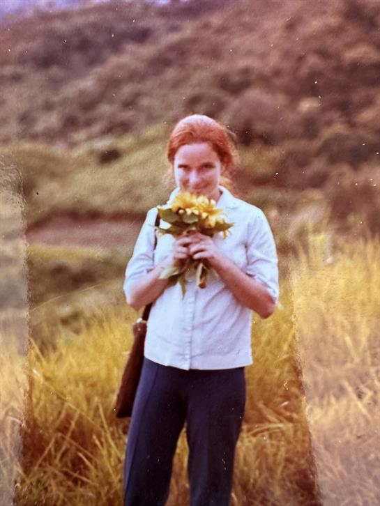 A woman with red hair is smiling while holding fresh flowers in a sunny grassy field.