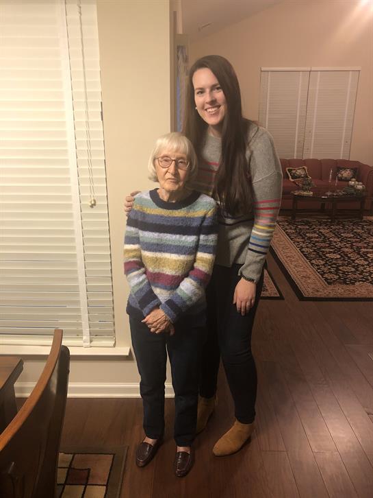 Two women smile together in a warm living room, showcasing their matching colorful sweaters.