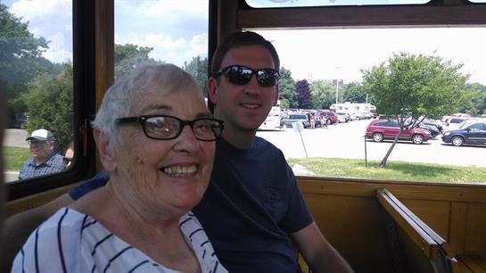 An elderly woman smiles widely with a young man on a joyful trolley ride.