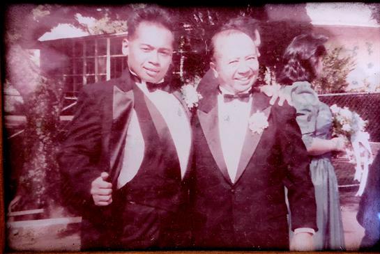 Two well-dressed men stand side by side, smiling warmly at a gathering, surrounded by greenery.