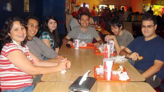 A group of six friends share laughter over a meal at a vibrant food court during the evening.
