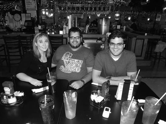 Three friends sit at a bar table with drinks, enjoying laughter and conversation at night.