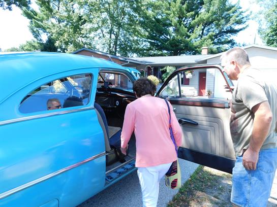 A woman approaches an old blue car while a man stands nearby offering assistance at a gathering.