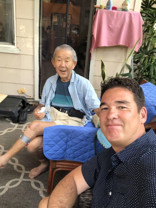 An older man and a younger man share a cheerful moment on a sunny porch while eating ice cream.