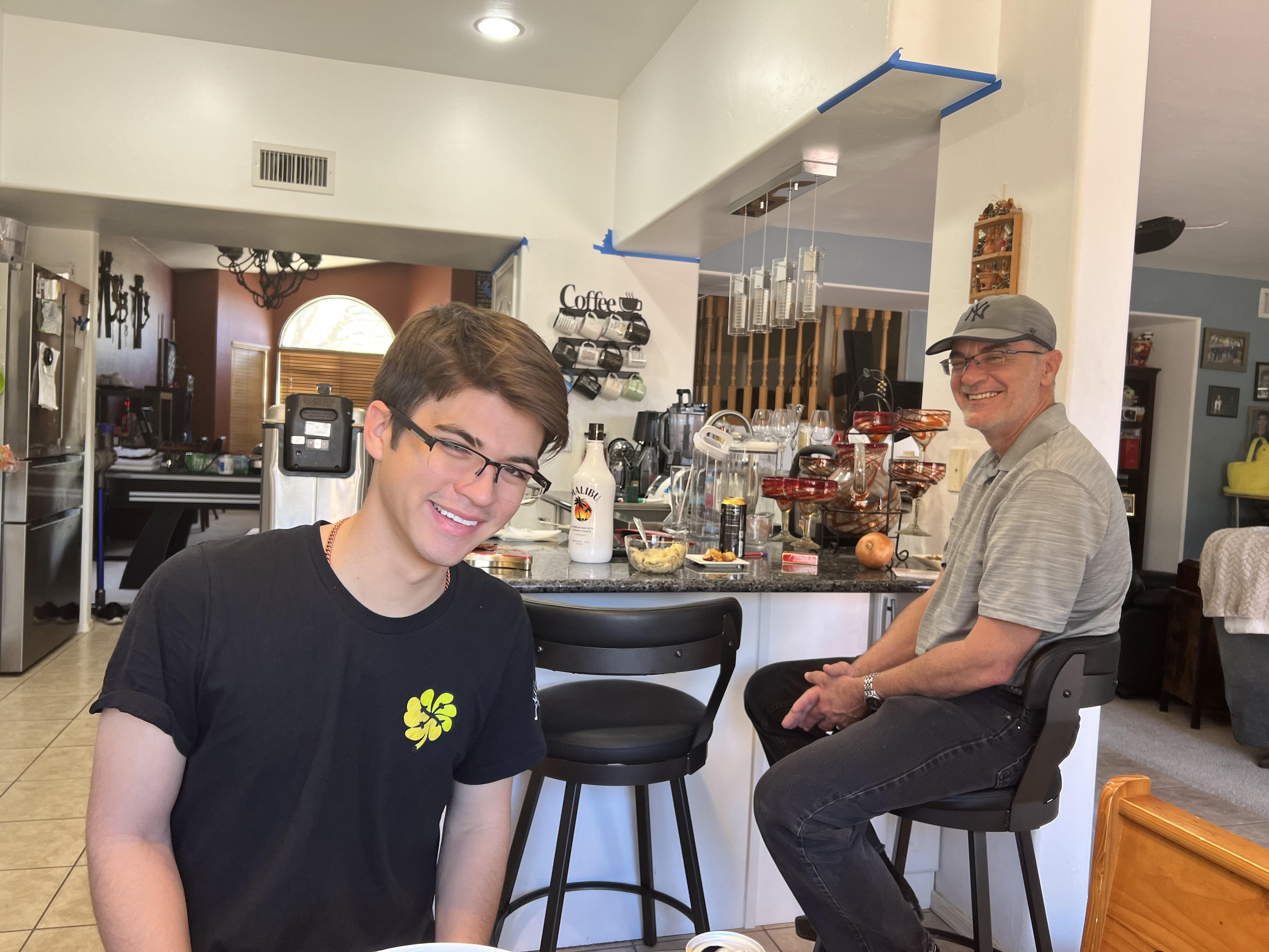 A young man and older man engage in friendly conversation at a kitchen counter filled with snacks.