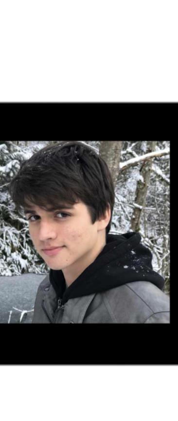 A boy with dark hair stands in a snowy forest, smiling amidst winter scenery.