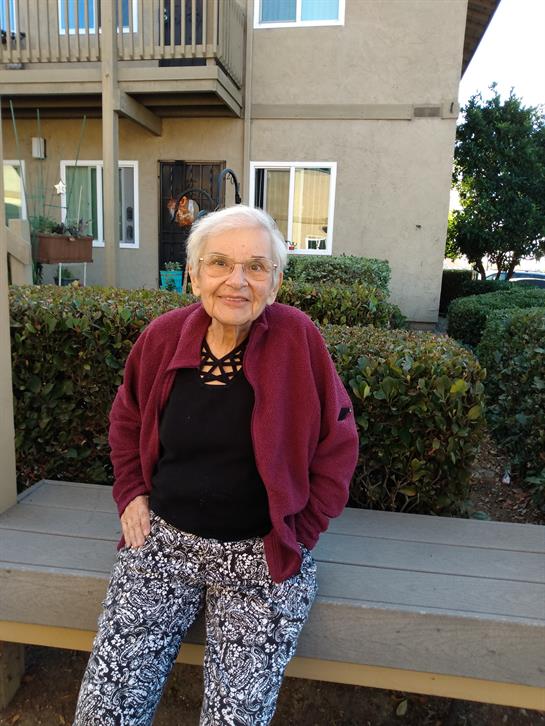 An elderly woman smiles while seated on a bench in a community garden area with greenery.
