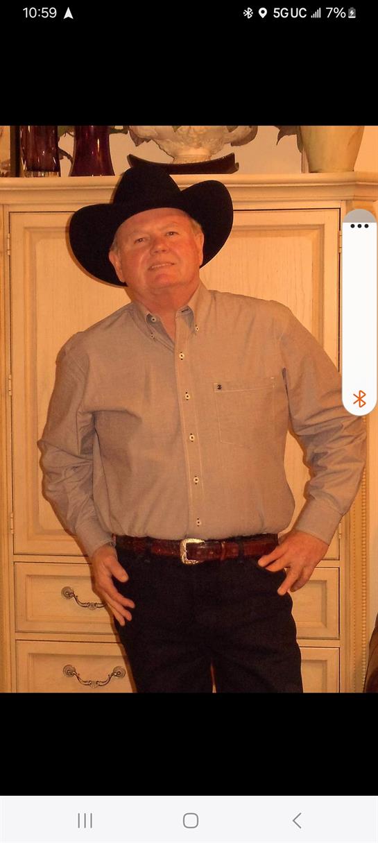 A man in a cowboy hat and button-up shirt stands with confidence, smiling indoors.