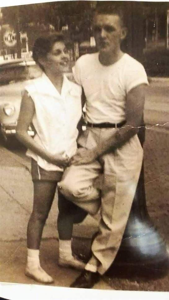 Two young adults stand closely together, smiling on a vibrant city street during the 1950s.