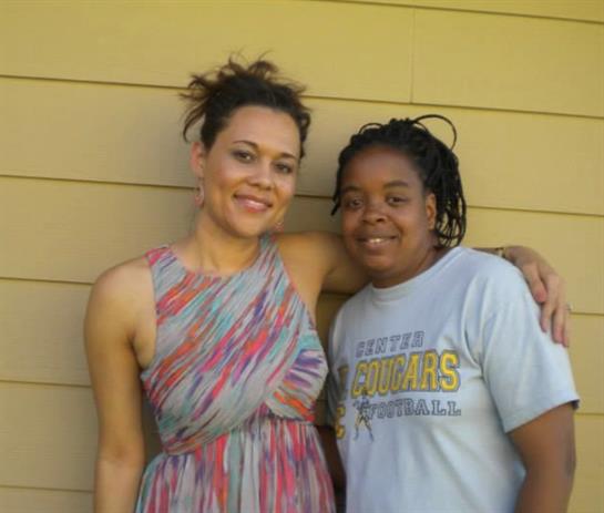 Two women stand closely side by side, smiling brightly in a charming outdoor location.