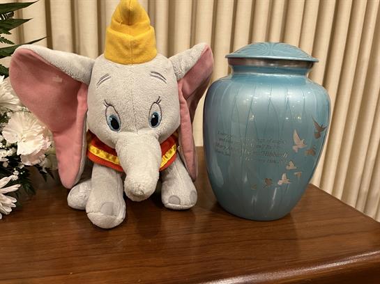 A soft gray elephant toy with big ears and a yellow hat sits beside a blue urn on a wooden table.