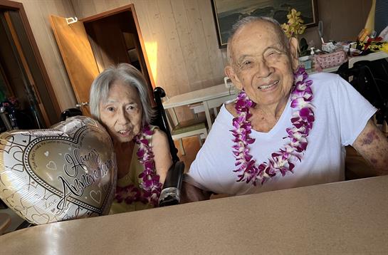 Elderly couple enjoys a joyful celebration with leis and a heart-shaped balloon in their home.