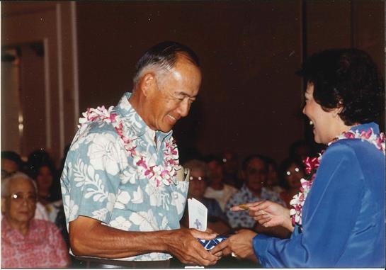 Two people share a joyful moment, exchanging gifts while surrounded by a festive crowd in Hawaii.