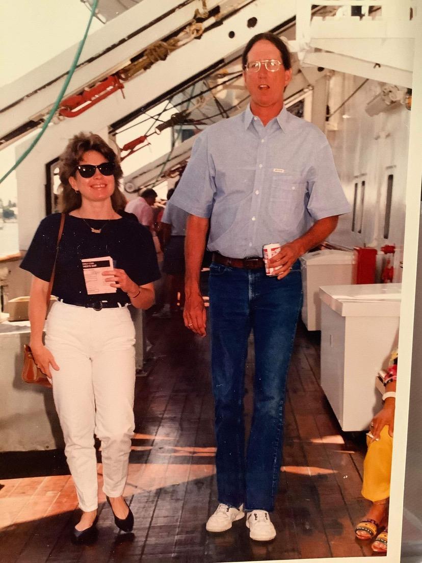A couple walks side by side on a ship's deck while holding tickets and a drink.