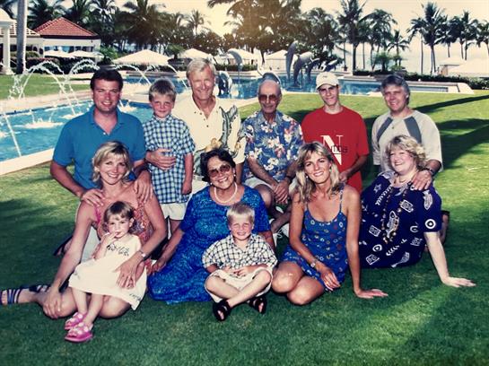 A family enjoys a joyful reunion outdoors near a pool, surrounded by palm trees and sunshine.