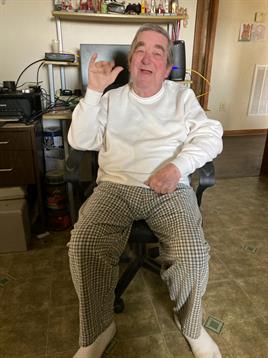 An elderly man is comfortably seated in a home office, smiling while gesturing.