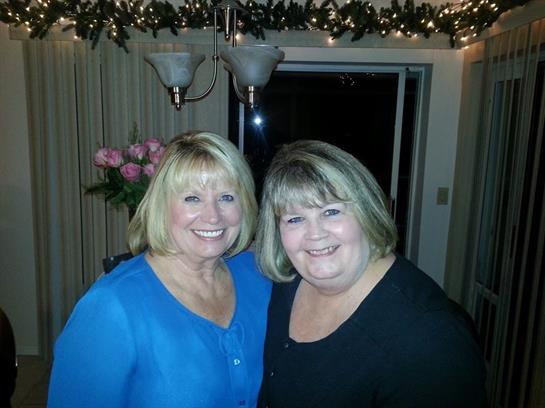 Two smiling women pose closely in a cozy indoor space adorned with lights and flowers.