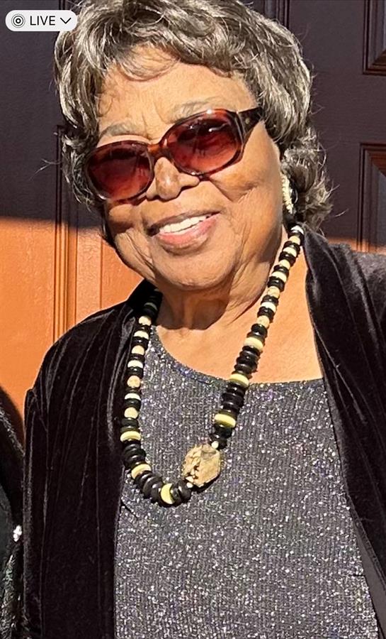 An elderly woman smiles in sunglasses and a necklace during an outdoor celebration.