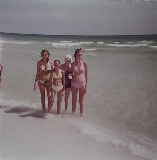 Four friends walk through shallow water at a beach, smiling and enjoying the summer sunshine.