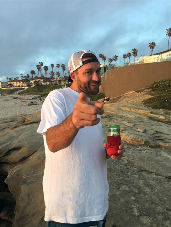 A man stands on a sandy beach at sunset, holding a drink and smiling, enjoying a carefree moment.