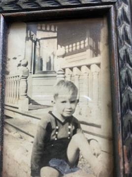 A young boy in suspenders sits on the steps of a vintage house, looking thoughtful and serene.