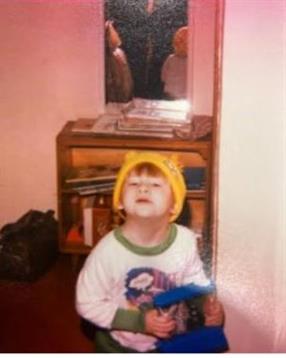 A young child with a playful expression wears a yellow hat and holds a toy gun while indoors.
