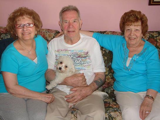 Three friends smile while sitting on a sofa, one holding a small dog, creating a warm atmosphere.