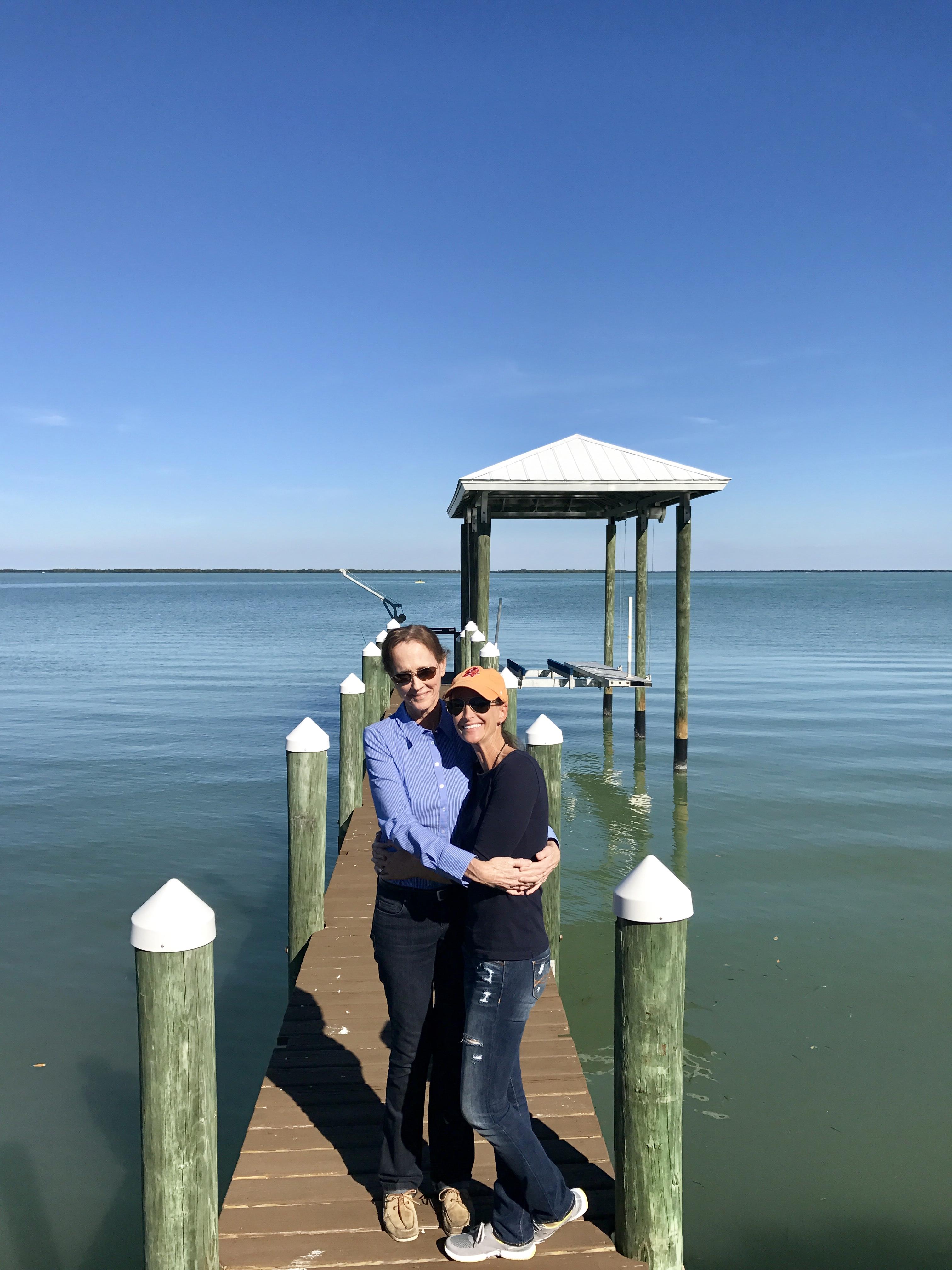 Two people embrace on a dock overlooking calm waters under a clear blue sky.