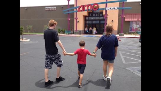 Three family members holding hands stroll toward a cinema in bright sunlight.