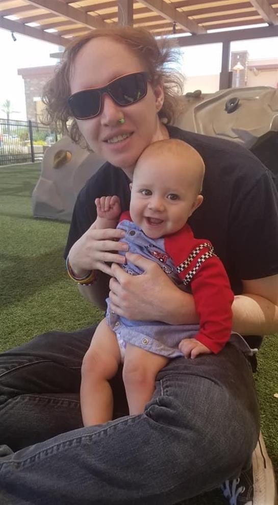 A baby sits on an adult's lap, both smiling and playing together in a grassy play area.