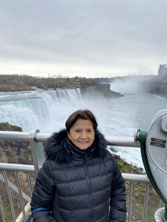 A woman stands near the railing, smiling with Niagara Falls in the background on a cloudy day.