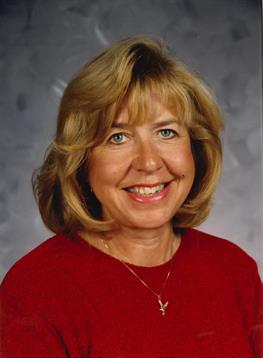 A blonde woman smiles warmly in a red sweater on a grey background.
